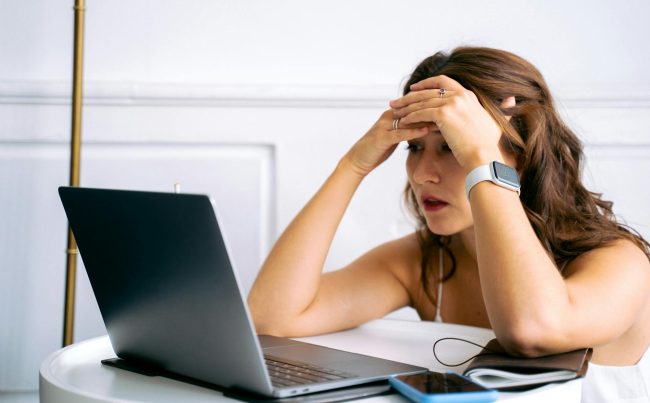 Dziewczyna siedzi przy biurku przed laptopem, trzyma głowę w dłoniach i wygląda na zmartwioną lub zestresowaną; obok leży smartfon i notes.