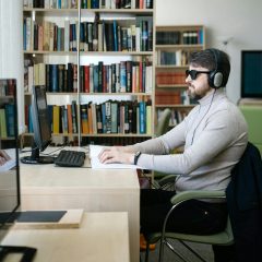 Niewidomy mężczyzna w ciemnych okularach czyta książkę w alfabecie Braille'a przy biurku, mając na uszach słuchawki. W tle widać komputer i regały z książkami.