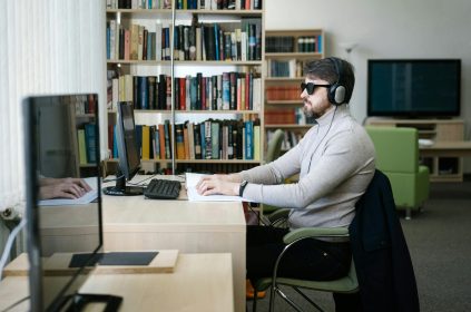 Niewidomy mężczyzna w ciemnych okularach czyta książkę w alfabecie Braille'a przy biurku, mając na uszach słuchawki. W tle widać komputer i regały z książkami.