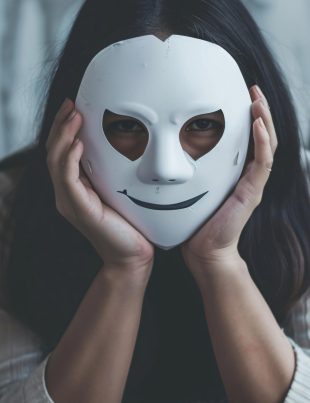 Kobieta trzymająca białą maskę z uśmiechem, zasłaniającą część twarzy — obraz symbolizujący ukrywanie prawdziwej tożsamości w internecie.