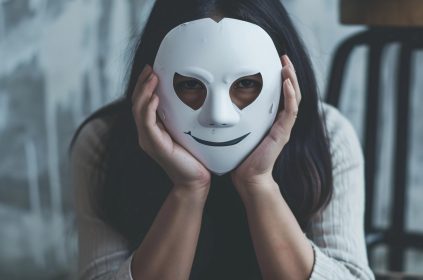 Kobieta trzymająca białą maskę z uśmiechem, zasłaniającą część twarzy — obraz symbolizujący ukrywanie prawdziwej tożsamości w internecie.