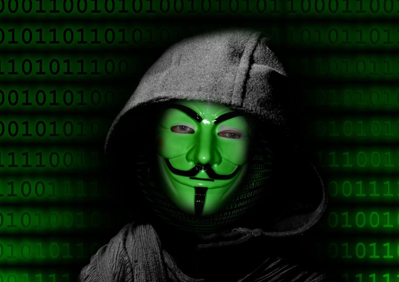 Osoba w zielonej masce Guy Fawkes na tle kodu binarnego, przedstawiająca cyberprzestępczość i hakerstwo.