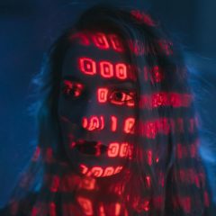 Twarz kobiety oświetlona czerwonym światłem z wzorami binarnych cyfr, symbolizująca zależność człowieka od technologii i danych.