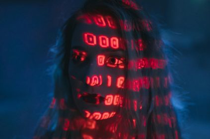 Twarz kobiety oświetlona czerwonym światłem z wzorami binarnych cyfr, symbolizująca zależność człowieka od technologii i danych.