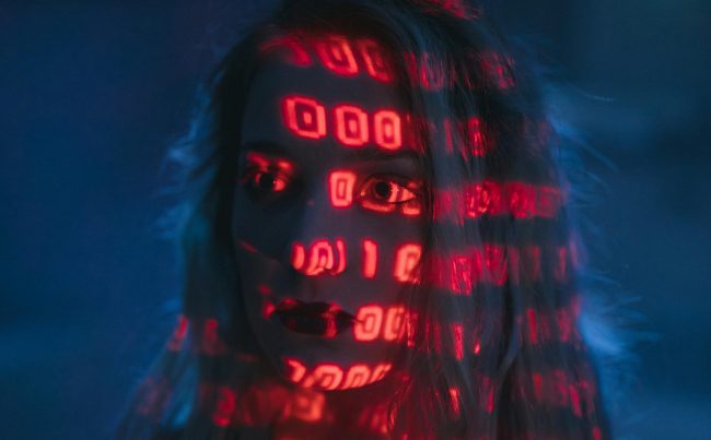 Twarz kobiety oświetlona czerwonym światłem z wzorami binarnych cyfr, symbolizująca zależność człowieka od technologii i danych.